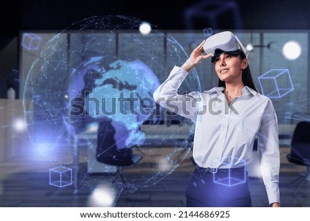 Attractive happy young european businesswoman using VR glasses with creative glowing polygonal blue globe metaverse on blurry office workplace background. Augmented reality and future concept. 
