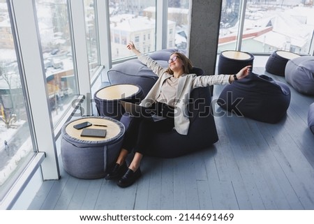 Successful young woman in a jacket smiling and working on a laptop in a modern office with large windows. Rest after work. Remote work