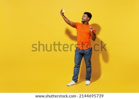 Full body young man of African American ethnicity 20s wear orange t-shirt doing selfie shot on mobile cell phone post photo on social network show v-sign isolated on plain yellow background studio.