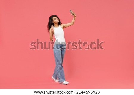 Full length young friendly african american woman in white tank shirt do selfie shot on mobile phone show victory v-sign gesture isolated on pink background studio portrait. People lifestyle concept