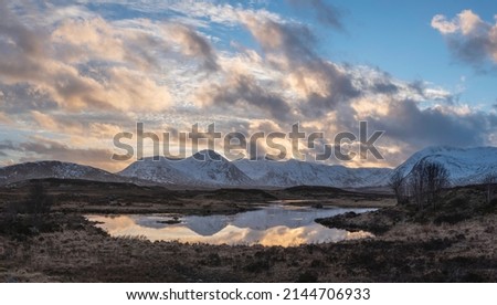 Stunning Winter panorama landscape image of mountain range viewed from Loch Ba in Scottish Highlands with dramatic clouds overhead