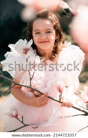 Beautiful smiling kid girl 4-5 year old wear stylish princess dress smell magnolia flower over blooming tree in garden outdoor. Spring season. Childhood. Springtime. Happy child with floral nature. 
