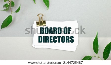 On a gray background - green leaves of the plant and a white card under a gold clip with the text BOARD OF DIRECTORS. Minimalist.