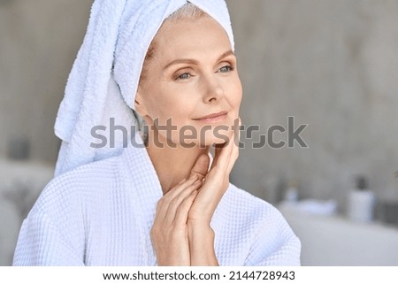 Happy beautiful middle aged woman wearing bathrobe and white towel with perfect complexion touching face looking away in bathroom. Advertising of skin care spa wellness concept. Closeup portrait. Royalty-Free Stock Photo #2144728943