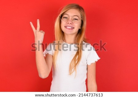 blonde little kid girl wearing white t-shirt over red background smiling and looking friendly, showing number two or second with hand forward, counting down