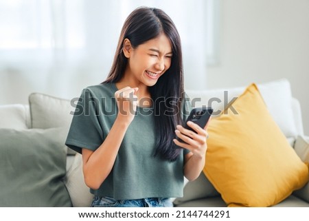 Excited young asian woman hold phone chatting in dating app feel happy sit on sofa at home, Closeup joyful female reading good news on phone. Surprised lady celebrating victory on phone Royalty-Free Stock Photo #2144744291