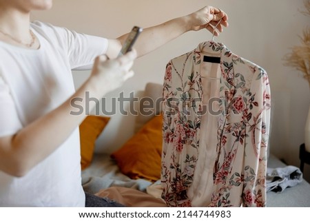 Young woman holding smartphone and taking photo of her old clothes to sell them online. Selling on website, e-commerce. Reuse, second-hand concept. Conscious consumer, sustainable lifestyle.