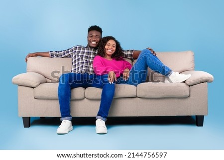 Happy millennial black couple relaxing on sofa, smiling at camera on blue studio background, full length. Young boyfriend and girlfriend resting on couch, having break together on free time Royalty-Free Stock Photo #2144756597