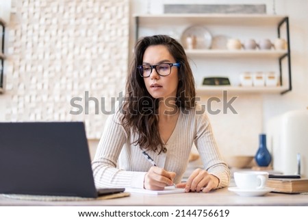 Freelance, remote manager, home accounting and tax payment. Woman in glasses making notes at kitchen table with laptop, studying or working online