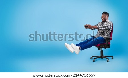 Excited young black guy with joystick sitting on chair, playing video game on blue background, banner with copy space. Happy male gamer with controller participating in online gaming championship