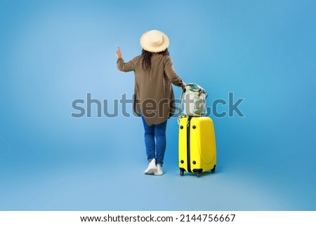 Back view of young black woman with luggage pointing, choosing tourist tour on blue studio background, copy space for travel offer or advertisement. Hot offer, summer vacation concept