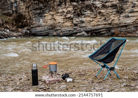 Tourist parking near the river, camping on the shore of the reservoir, kitchen for cooking on a hike, folding chair, thermos with tea, pot, dishes on the ground. High quality photo