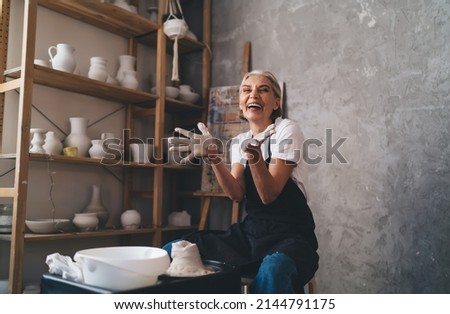 Cheerful caucasian senior female sculptor with opened hands while she making clay pot on pottery wheel. Idea of small business and entrepreneurship. Home hobby, entertainment and leisure. Art studio