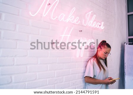 Hipster girl in eyeglasses using cellphone for chatting near illuminated banner logo lettering with motivation phrase, female millennial in spectacles creating publication near colorful neon text