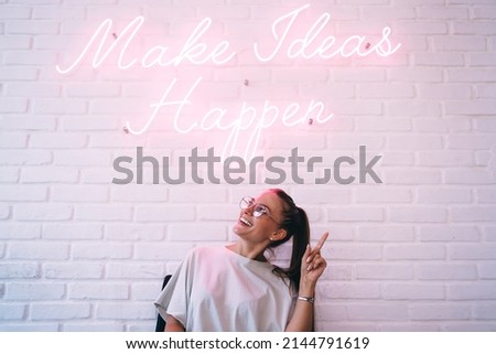 Happy millennial hipster girl in trendy spectacles smiling near icon slogan with inspirational and motivational quote text - LED illumination at bricked wall, cheerful teenager near font neon message