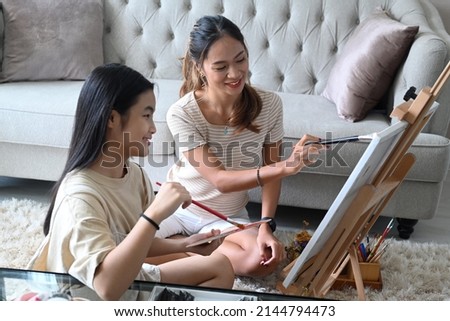Smiling asian woman and her young sister sitting in front of easel and painting picture on canvas.