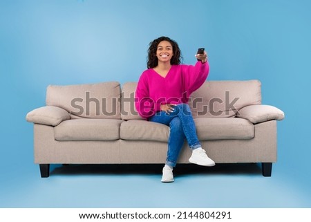 Young African American lady with remote control sitting on couch and watching TV on blue studio background, copy space. Millennial woman with television controller switching channels Royalty-Free Stock Photo #2144804291
