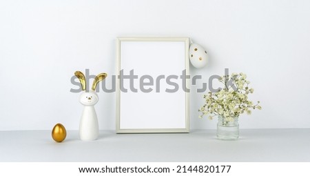 Easter decor. Frame mock up for your text. Eggs and fun rabbit with golden ears. Flowers on background of white table and wall