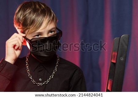 Portrait of a stylish teenager 13 years old with a fashionable hairstyle in black sunglasses, a protective mask and with a chain around his neck on a purple background. Selective focus. Close-up