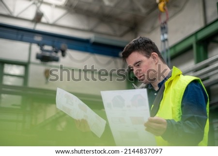 Young man with Down syndrome looking at blueprints when working in industrial factory, social integration concept. Royalty-Free Stock Photo #2144837097