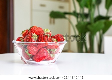 A glass bowl of juicy strawberries on white table of living room interior, cupboards, drawers, and Giant White Bird of Paradise plant (Strelitzia nicolai) out of focus in background, healthy lifestyle