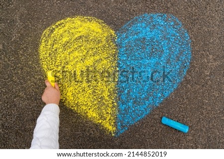 A child's hand draws a heart with crayons on the pavement with the patriotic national Ukrainian colors of yellow and blue. Stop the war in Ukraine. High quality photo
