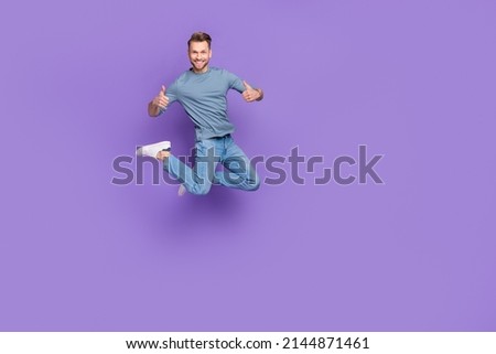 Photo of pretty confident guy wear grey shirt jumping high thumbs up empty space isolated violet color background