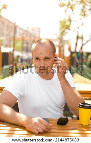 The person inserts wireless headphones into the ear. A man is sitting in a cafe with a cup of coffee and establishes a connection between headphones and a phone to listen to music