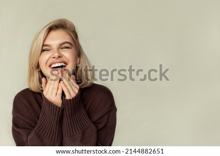 a shot of a joyful blonde with a short haircut, a young cute woman in a jumper laughs sincerely on a beige studio wall. Concept of happiness and positive emotions