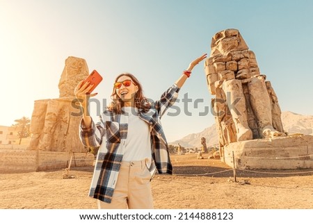 Travel blogger girl takes selfie pictures on a smartphone at the famous two Colossi of Memnon - massive ruined statues of the Pharaoh Amenhotep III. World tourism attractions