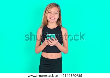 Pleased blonde little kid girl wearing pink sport clothes over green background using self phone and looking and winking at the camera. Flirt and coquettish concept.