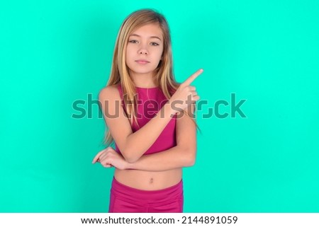 blonde little kid girl wearing pink sport clothes over green background smiling broadly at camera, pointing fingers away, showing something interesting and exciting.