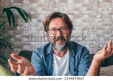 Bearded middle aged man in casual clothes and glasses gesticulating and talking to camera while sitting on sofa against brick wall in daytime at home Royalty-Free Stock Photo #2144893867