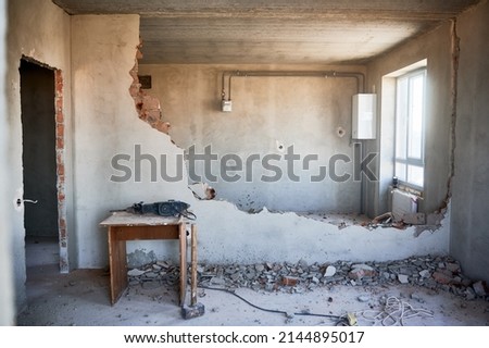 Housing redevelopment. Small table with perforator, sledgehammer placed on floor at partially destroyed inner wall. On backdrop empty walls, window, gas boiler, meter and pipes connecting them. Royalty-Free Stock Photo #2144895017
