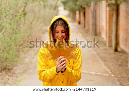High angle view of happy woman with raincoat holding maple tree leaves. Horizontal mid waist view of woman gathering fallen leaves in yellow hoodie outdoors. People and nature backgrounds.