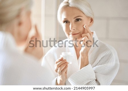 Attractive mid age older adult 50 years old blonde woman wears bathrobe in bathroom applying nourishing antiage face skin care cream treatment, looking at mirror doing daily morning beauty routine. Royalty-Free Stock Photo #2144905997