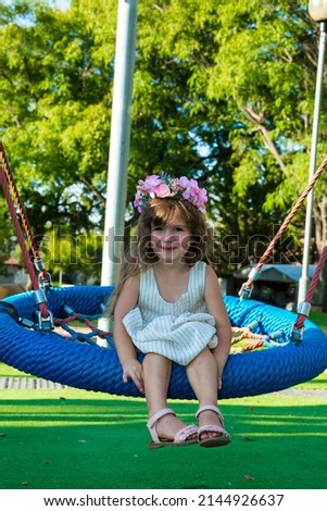 Warm summer day in the children's park. Beautiful little girl with long blond curls in a flower wreath on a children's swing