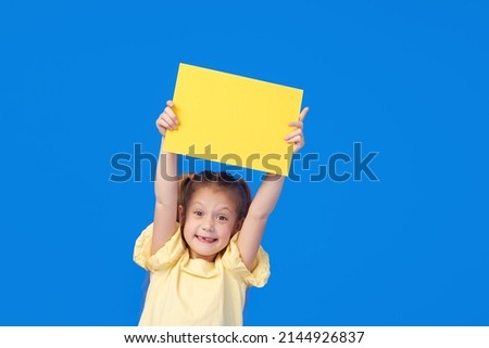 A cute little girl is holding a yellow sign for an inscription on a blue background. Funny face.
