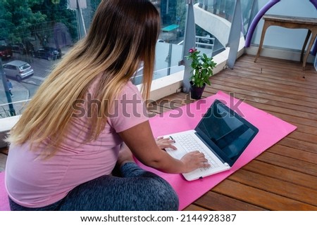 rear view of pregnant unrecognizable caucasian young woman sitting on pink yoga mat at home typing on laptop keyboard, practicing yoga and meditation online.