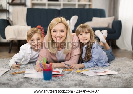 Little sister, brother and their mother playing on warm floor with underfloor heating in living room. Cute girl, boy and mom drawing with colorful pencils, enjoying leisure time at home.
