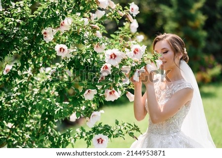 Close view of cute girl with natural makeup and veil on her hair, wearing in wedding dress, which adorned by lace and beads, holding and smelling flowers which growing on bush, closing eyes and relax Royalty-Free Stock Photo #2144953871