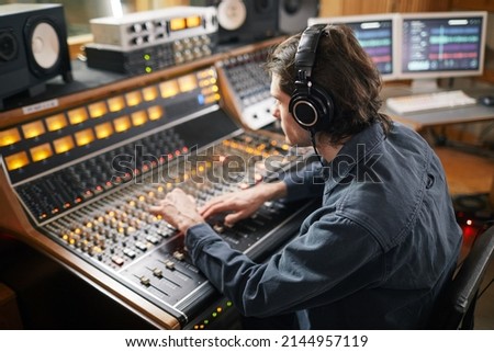Portrait of young man wearing headphones and operating buttons and toggles at digital audio workstation in recording studio, music production concept Royalty-Free Stock Photo #2144957119