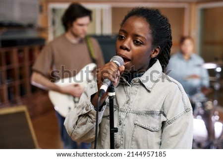 Waist up portrait of young black woman singing to microphone while recording music with band in professional studio