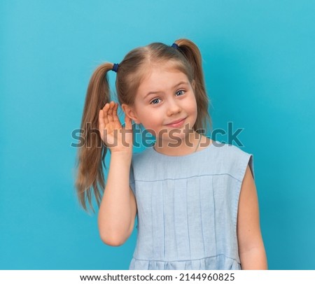 Positive smiling little girl holds hand near the ear listening, isolated on blue background.