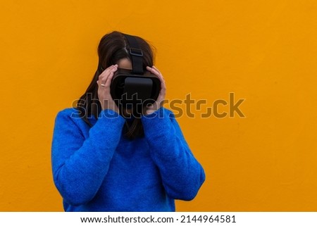 portrait of a young woman with a look of fear or bewilderment wearing virtual reality goggles on a yellow background