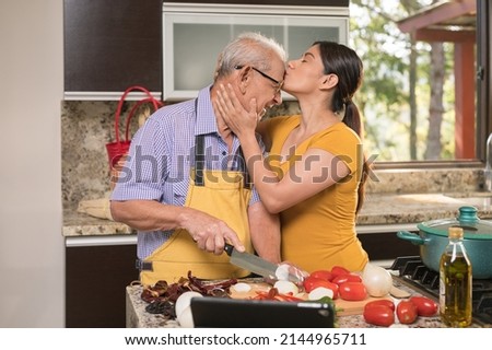 Daughter affectionately kisses her father on the forehead while he prepares the food.