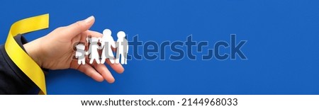 Helping hand holds Ukrainian family, white paper silhouette. Flat lay, top view with yellow ribbon on blue background. Panorama, volunteer help to refugees from Ukraine. Copy-space, panoramic banner.