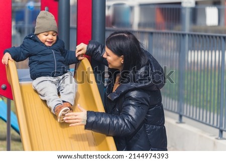 dark-haired woman with a long black coat looking at a sweet little boy who is sliding down on a plastic playground outdoors cowboy shot. High quality photo Royalty-Free Stock Photo #2144974393