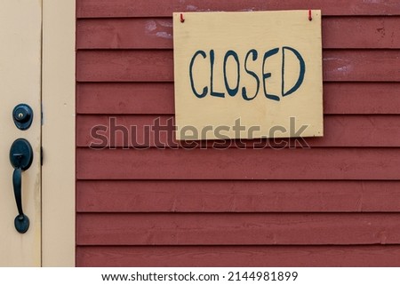 A yellow wooden closed sign with black letters on a red wooden building. The narrow cape cod clapboard wall has a beige colored door with a black metal handle and deadbolt lock. 