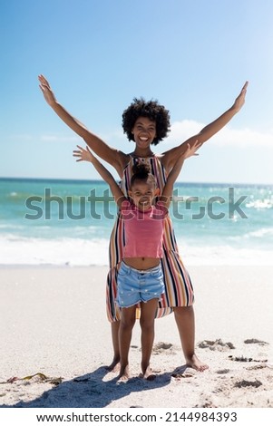Full length portrait of happy african american mother and daughter with arms raised at beach. unaltered, family, lifestyle, togetherness, enjoyment and holiday concept.